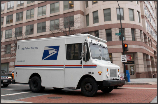 A USPS delivery truck driving through an urban intersection.